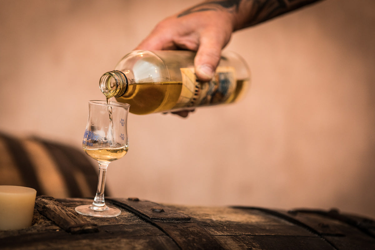 Nahaufnahme von Hoffer Alter Schnaps, der aus einer Flasche in ein kleines Verkostungsglas auf einem Holzfass eingeschenkt wird, in warmer, rustikaler Atmosphäre.