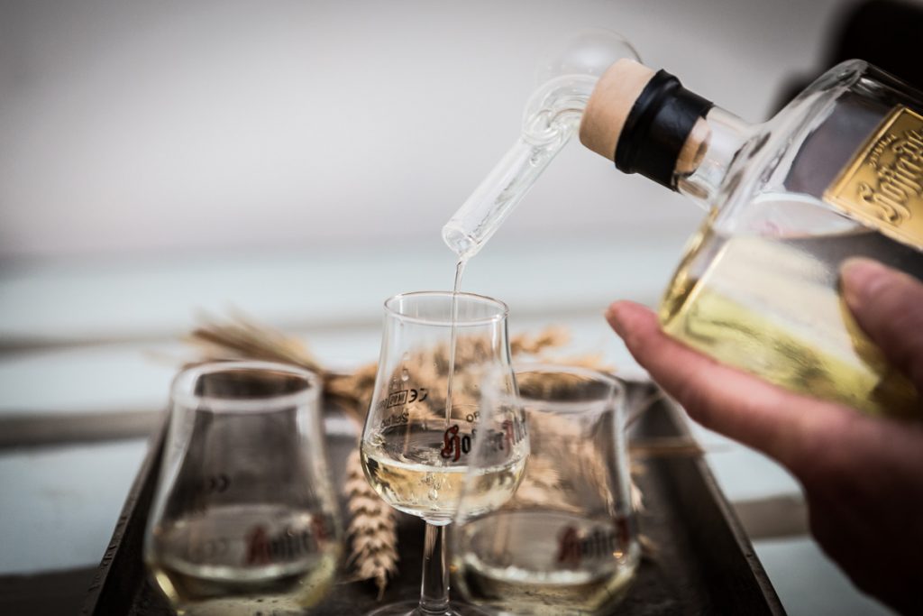 Nahaufnahme von Hoffer Alter Schnaps, der aus einer Glasflasche in ein kleines Verkostungsglas eingeschenkt wird, mit zwei weiteren Gläsern und Weizen im Hintergrund.